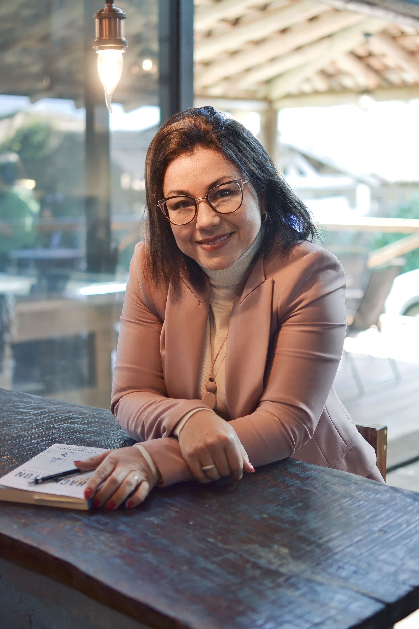 Gislaine Lavinia, sorrindo em um ambiente acolhedor, com um livro sobre a mesa.